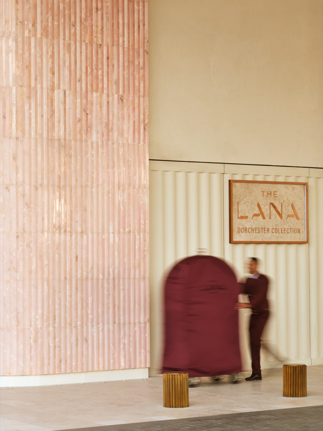 A doorman at The Lana, Dorchester Collection, moving a covered luggage cart through a softly lit arrival hall